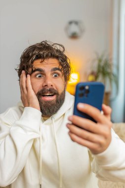 Akıllı telefon kullanan genç Hintli sakallı bir adam, iyi haber mesajı alıyor, piyango zaferini kutlarken şok oluyor evdeki kanepede oturma odasında büyük ikramiyeyi kazanıyor. Yaşam Tarzları.