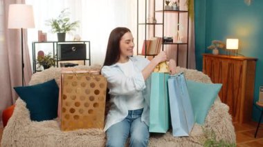 Genç bir kadın alışveriş çantasından aldığı yeni kıyafetleri mutlulukla açıyor. Kanepede oturan beyaz bir kız alışveriş sonrası heyecanlı, şık bir moda ürününden memnun.
