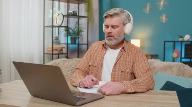Beyaz, olgun bir adam evde oturmuş kulaklıklı bir dizüstü bilgisayardan podcast ya da online yayın dinliyor. Adam not defterine not tutuyor, içerikle ilgileniyor, eğitimi öğreniyor.