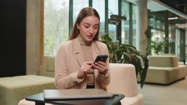 Mutlu iş kadını cep telefonu daktilosu kullanıyor, başarılı zaferi kutluyor, piyango büyük ikramiyesi kazanıyor, oyun oynuyor, iyi haberler, modern ofis lobisinde zafer kazanıyor.