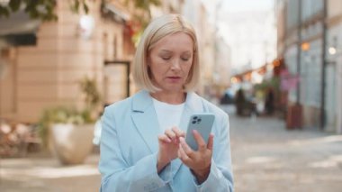 Şehir caddesinde duran mutlu bir iş kadınının portresi. Resmi takım elbiseli, gülümseyen beyaz kadın işveren akıllı telefonu kullanıyor ve kameraya bakıyor. Pozitif kadın yöneticinin vesikalığı..