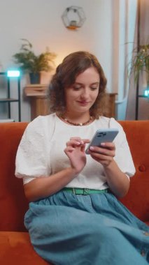 Oturma odasında gülümseyen cep telefonuyla kanepede oturan genç bir kadının portresi. Akıllı telefon sosyal medya uygulamalarında internetten film izleyen kız mesajları paylaşıyor.