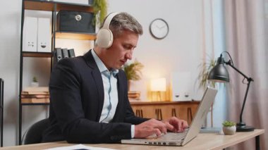 Genç ve mutlu bir iş adamı ofis masasında oturmuş kulaklıkla en sevdiği müziği dinliyor. Modern odada dizüstü bilgisayarla çalışan resmi takım elbiseli, güler yüzlü, serbest çalışan beyaz adam..