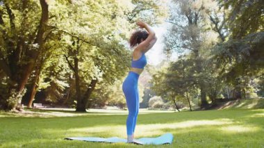 Spor kıyafetli beyaz kız parkta yoga minderinde esneme egzersizleri yapıyor. Sporcu kadın çimde öne eğilip pratik yapıyor. Sağlıklı yaşam tarzını temsil eden atletik kadın..