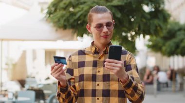 Genç adam para transfer ederken kredi kartı akıllı telefonu kullanıyor, online alışveriş yapıyor, yemek siparişi veriyor, otel odası ayırtıyor. Şehir merkezinin güneşli sokaklarında mutlu bir adam. Yaşam Tarzları.