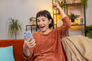 Evdeki kanepede oturmuş akıllı telefon kazanan piyangoyu karıştıran genç bir kadın. Heyecanlı Kafkasyalı kız dairede zafer kutlaması yapıyor yumruklarını sıkıyor. Mutlu bayan çevrimiçi oyun oynuyor