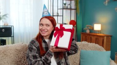 Evde parti şapkası takan genç bir kadın güzel bir sürpriz tepkiyle doğum günü hediyesi veriyor ve gülümsüyor. Kafkasyalı kızıl saçlı kız kanepenin üzerinde sevinç ve duygusal zevkle kutlama anının tadını çıkarıyor.