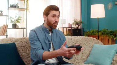 Genç adam evinde kanepede oturuyor, oyun konsolunu kavrıyor, yoğun bir şekilde odaklanarak ve heyecanla oyun oynuyor. Kafkasyalı adam oyun oynamaya duygusal tepki veriyor, sanal dünyaya dalmış, kapalı mekanlarda.