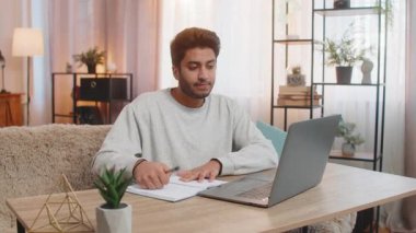 Masada oturan genç Hintli adam dizüstü bilgisayarla çevrimiçi sınıfa katılır, dikkatle ders dinler, not defterine notlar alır. Evdeki İspanyol adam, ya kendi işine odaklanmış ya da yakınlardaki aktivitelere odaklanmış..