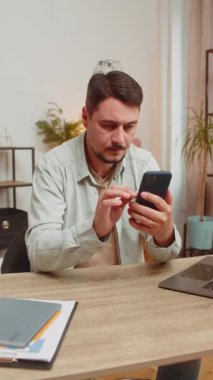 Ofiste gülümseyen cep telefonuyla masada oturan mutlu beyaz bir adam. Serbest çalışan biri, internetten akıllı telefon sosyal medya uygulamalarından mesaj yolluyor. Dikey.