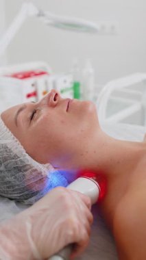 Kozmetik uzmanı, kadını yüz ve boyun bölgesinin kaldırma prosedürü, kolajen maskesi yapar. Güzellik uzmanı vakumlu yüz masajı terapisi için elektrikli ekipman kullanıyor. Cilt bakımı, güzellik tedavisi. Dikey çekim