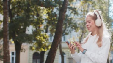 Kablosuz kulaklıklı mutlu, gevşemiş kadın turist dışarıda dans eden akıllı telefondan disko müziği dinliyor. Şehir caddesinde günlük giysiler içinde yürüyen genç kız. Kasaba yaşam tarzları.