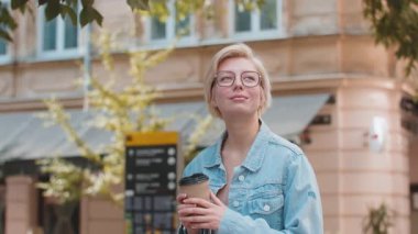 Kafkasyalı genç, mutlu sarışın kadın sabah kahvesinin tadını çıkarıyor sıcak bir şeyler içiyor ve dışarıda gülümsüyor. Dinleniyorum, ara veriyorum. Şehir merkezi caddesinde yürüyen ve kahve içen bir kız. Kasaba yaşam tarzları.