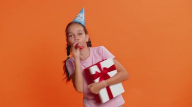 Doğum günü kutlaması şapkası takmış, başarıyı kutlayan pipo üfleyen çocuk hediye kutusu tutuyor. Zaferi kazanıyor. Yıldönümü partisindeki heyecanlı çocuklar turuncu arka planda izole edilmiş.