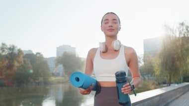 Elinde hasırla genç ve mutlu sporcu kadın parktaki açık hava eğitimine gidiyor. Aktif giyimli, kulaklık takan ve su şişesi kullanan iyimser gülümseyen kız jimnastik eğitimine gidiyor..