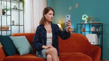 Genç kadın blogcu akıllı telefondan selfie çekiyor, abonelerle çevrimiçi video görüşmesi yapıyor, sosyal medya için hikayeler kaydediyor. Evde oturan kız kanepede oturuyor. Yaşam Tarzları.