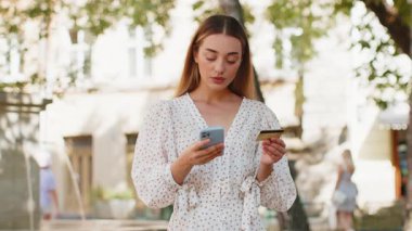 Kafkasyalı genç bir kadın kredi kartı akıllı telefonu kullanıyor. Para transfer ederken, online alışveriş yaparken, yemek siparişi verirken, açık havada otel odasında rezervasyon yaparken. Şehir caddesinde giyinmiş mutlu kız.