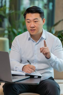 Genç Çinli iş adamı, otel lobisindeki internet video dersi veya ofis toplantısına katılırken not alıyor ya da not alıyor. Web kamerasına bakan gömlek destek servislerinde kendine güvenen bir yönetici