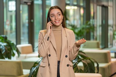 Resmi görevde gülümseyen genç bir iş kadını, modern ofis lobisinde cep telefonuyla konuşuyor. Ulak sohbet uygulamaları kullanarak akıllı telefonu tutan profesyonel yönetici kadın. İstihdam, meslek.