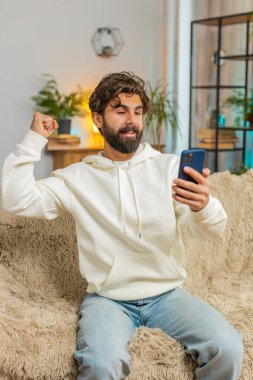 Mutlu genç adam akıllı telefon daktilo kullanarak evet diyor, başarıyı kutluyor, piyango hedefini kazanıyor olumlu haberler, zafer. Evde koltukta oturan sakallı adam..