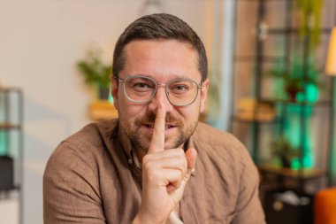 Sessiz olun lütfen. Kafkasyalı genç işadamı işaret parmağını dudaklarına bastırıyor. Ofis masasında sessiz jest işareti yapıyor. Oturma odasında dizüstü bilgisayarlı yetişkin bir adam..