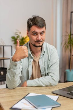 Gibi. Mutlu, heyecanlı, beyaz bir adam kameraya bakıp olumlu bir işaret veriyor. İyi haber, olumlu geri bildirim. Genç adam ofis masasında oturuyor. Dikey.