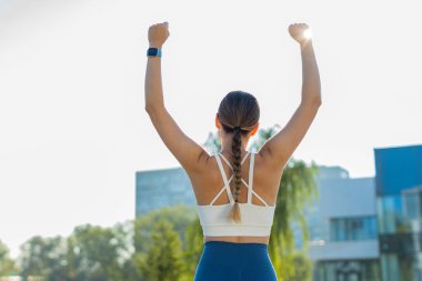 Güneşli bir günde şehir parkında koşu yaptıktan sonra başarıyı kutlayan heyecanlı beyaz atletik kadın koşucusunun arka görüntüsü. Koşu şampiyonu maratonu kazanıyor. Aktif sporcu kadın motivasyonu.
