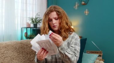 Evde elinde faturalar ve makbuzlar tutan genç bir kadın. Kafası elinde. Masraflar yüzünden üzgün ve stresli görünüyor. Kanepede oturan beyaz saçlı kız duygusal baskı, maddi sıkıntı ve hayal kırıklığı hissediyor.