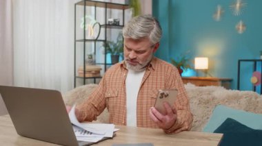 Beyaz, olgun bir adam evde, dizüstü bilgisayar ve akıllı telefon kullanıyor, mali raporları inceliyor. Orta yaşlı bir adam yorgun görünüyor, gözlerini ovuşturuyor ve finansal sorunlara özenle çözüm bulmaya çalışıyor.