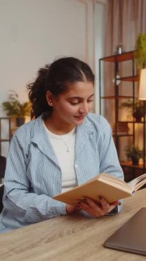 Hint genç kadın kitap okuyor, sayfalarca gülümsüyor edebiyattan zevk alıyor, masa başında dinleniyor. Huzurlu, neşeli bir kızın portresi. Oturma odasında dinleniyor. Dikey.