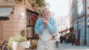 Dışarıda dikilen heyecanlı kadın sevinçten havalara uçuyor zaferi kazanmanın hazzını hissederek başarı evet işareti yapıyor. Şehir caddesinde sarı saçlı bayan gezgin..