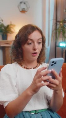 Akıllı telefon daktilosu kullanan mutlu ve heyecanlı genç bir kadın büyük bir zafer kazandı. İyi haberler zaferi kutlar. Cep telefonlu, evinde kanepede oturan Kafkasyalı hayret verici kız. Vay canına.