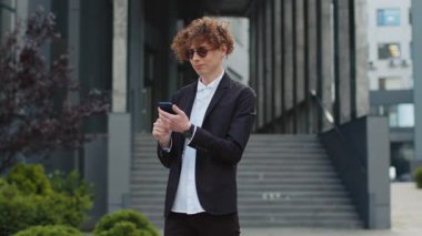 Kafkasyalı mutlu iş adamı akıllı telefon daktilosu kullanıyor e-posta mesajlarına cevap veriyor açık havada internetteki mobil uygulamalarla sohbet ediyor. Şehir merkezinde takım elbiseli kıvırcık saçlı yakışıklı bir adam. İş adamları