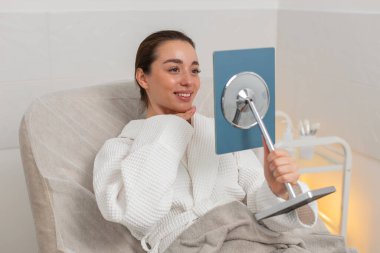 Beyaz kız güzellik kliniğinde oturuyor ve kozmetoloji ameliyatından sonra aynada yüzünü kontrol ederken gülümsüyor. Hintli genç kadın tedaviden memnun, sağlıklı ve taze deri sonuçlarının tadını çıkarıyor..