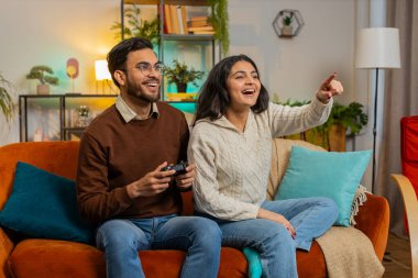 Oyun kumandası kullanan neşeli İspanyol çift oturma odasındaki kanepede oturup eğleniyor. Çeşitli aileler hafta sonu evlerinde online maç kazanmanın keyfini çıkarıyor.