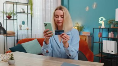 Kredi kartı ve akıllı telefon kullanan genç bir kadın para transferi yaparken internetten alışveriş yapıyor ev ofisinin masasında oturuyor. Çevrimiçi uzaktan kumanda işinde çalışan neşeli bir kız..