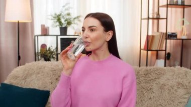 Sıcak havadan sonra susamış genç bir kadın saf filtrelenmiş su içiyor. Kanepede oturan beyaz bir kız sağlıklı yaşam tarzını, susuzluğunu iyi alışkanlıklarını tatmin ve tazelenmiş duygusal duygularını destekliyor.