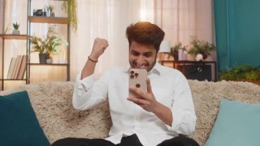 Hintli adam evinde koltukta gülümsüyor piyangoyu kazandıktan sonra heyecanlı bir şekilde akıllı telefonda yazı yazıyor, mutlu hissediyor. Arap adam başarıyı kutluyor, neşeli, heyecanlı, parlak duygularla harika haberlere tepki veriyor.