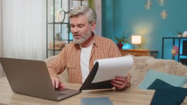 Beyaz, olgun bir adam evde oturmuş önemli belgeleri inceliyor ve dizüstü bilgisayar kullanıyor. Orta yaşlı bir adam başarılı bir işten aldığı memnuniyetle gülümser, verimlilik ve başarıyı gösterir.