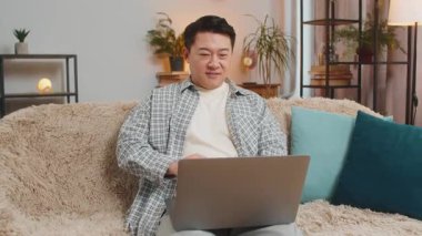 Oturma odasındaki işi bittikten sonra kanepeye oturmuş gülümseyen Çinli adam laptopu kapatıyor. Mutlu orta yaşlı adam evde online uzaktan kumanda işinde çalışıyor. Bilgisayarda E-öğrenme, internette gezinme.