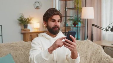 Mutlu Hintli adam akıllı telefon daktilo kullanarak evet diyor, zaferi kutluyor, piyango hedefini kazanıyor olumlu haberler, zafer. Evdeki kanepede oturan Arap adam..