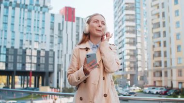 Kablosuz kulaklıklar takmış, dans eden, açık havada dans eden enerjik disko müziği dinleyen mutlu orta yaşlı iş kadını. Şehir merkezindeki caddede duran uzun montlu neşeli kız.