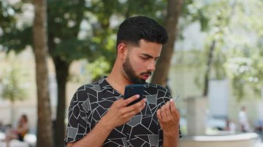 Hintli genç adam para transfer ederken kredi kartı akıllı telefonu kullanıyor, online alışveriş yapıyor, yemek siparişi veriyor, açık hava otel odası rezervasyonu yapıyor. Şehir sokağındaki İspanyol adam. Yaşam Tarzları.