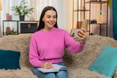 Evde kulaklık takan, cep telefonuyla eğitim videosu izleyen, dikkatle not alan genç bir kadın. Kanepede çalışan beyaz bir kız, internetten yeni bilgiler öğreniyor.