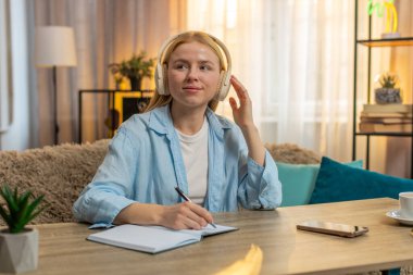 Genç bir kadın evde kulaklıkla çalışıyor, internetten meraklı bir şekilde ders dinliyor. Masa kayıtlarındaki beyaz kız notlar, günbatımında konfor ve beceriyi birleştirerek motivasyon,.