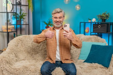 Olgun bir adam evdeki kanepede oturuyor, akıllı telefon tutuyor, baş parmağını hareket gibi kaldırıyor. Orta yaşlı adam sosyal medyada onayını gösteriyor, mutlu bir şekilde gülümsüyor, olumlu içeriğin tadını çıkarıyor iyi bir oyun uygulaması