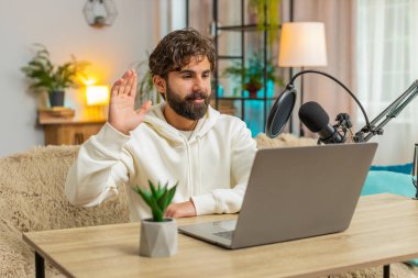 Mutlu Hintli blogcu erkek podcast 'i kanepeye koyup atölyeye Webinar veriyor. Genç İspanyol adam mikrofon ve dizüstü bilgisayarda konuşuyor, video görüşmesi yapıyor, evde oturmayı tartışıyor.