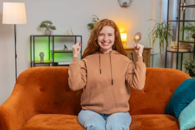 Mutlu genç bir kadın TV izlerken bağırıyor, başarıyı kutluyor, kazanıyor, gol başarısı iyi haber, piyango şansı, oturma odasında koltukta oturan zafer. Yetişkin kız evde çifte onay gösteriyor..