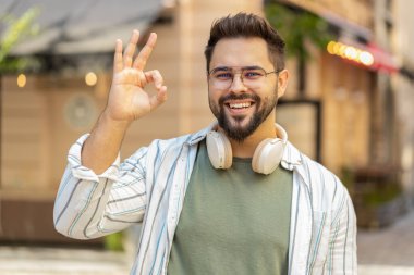 Gülümseyen genç milenyum çocuğunun el işaretini gösteren bir portresi. Beyaz tenli yakışıklı adam her şeyi gösteriyor. Güzel vücut dili. Gözlüklü turist kameraya gülümsüyor..