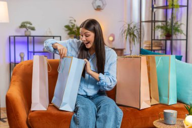 Tatmin olmuş Kafkasyalı genç bayan alışveriş kolik tüketicisi alışveriş merkezinde kağıt torbalarla alışveriş yaptıktan sonra eve geri döndü. Kız, kanepede otururken online siparişten paketler almış.